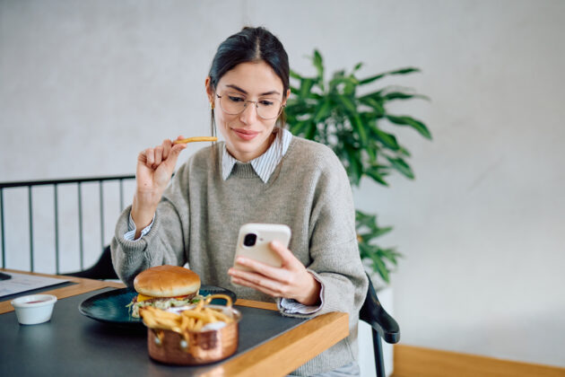 The picture shows a woman eating burger while looking at her phone